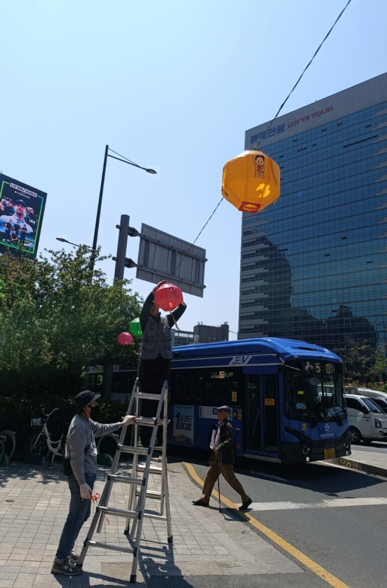 אדם עומד על סולם ותולה פנסים על חוט מעל רחוב עירוני, לצד אוטובוס כחול ובניין זכוכית מודרני, היוצרים אווירה של זכרון ותקווה בנוף העירוני. - דפנה פורמן, הרצאות, ייעוץ וטיפול רגשי במעברים וברילוקיישןש