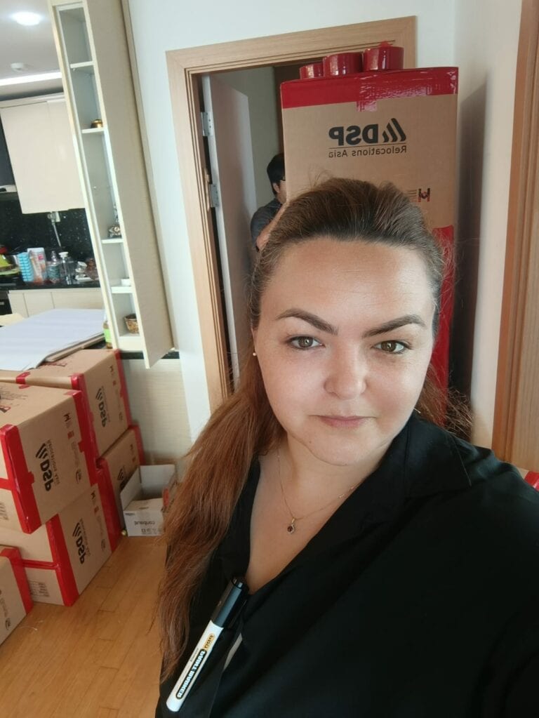 אישה מצלמת סלפי בחדר מלא בארגזים ובקופסאות עם הכיתוב "ASP". ברקע, ליד הדלת, נראית דמות חלקית של אדם נוסף, המבטא את רוח אריזת החיים. - דפנה פורמן, הרצאות, ייעוץ וטיפול רגשי במעברים וברילוקיישןש
