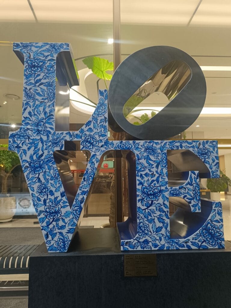 פסל המורכב מהמילה "LOVE" בדוגמת פרחים כחולים ולבנים מוצג בתוך המבנה, עם תאורה וצמחים ברקע, המסמל את האופן שבו אנו מתחברים לשנה באמצעות אמנות וחיבור. - דפנה פורמן, הרצאות, ייעוץ וטיפול רגשי במעברים וברילוקיישןש