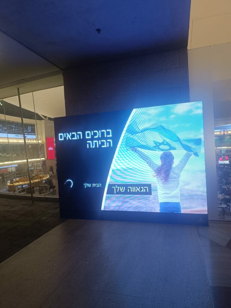 שלט דיגיטלי במרחב ציבורי מקורה מציג אדם מחזיק דגל ישראל, עם הכיתוב בעברית "וולקם טו איזראל", המברך את האנשים על חזרתם הביתה לישראל. - דפנה פורמן, הרצאות, ייעוץ וטיפול רגשי במעברים וברילוקיישןש