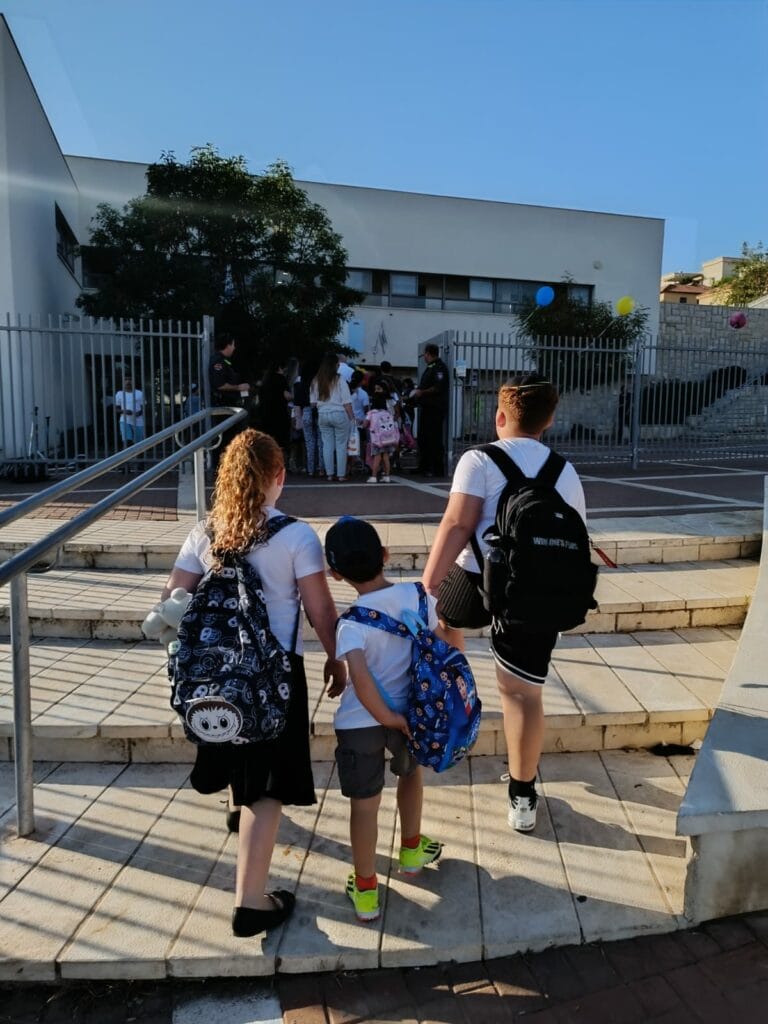 שלושה ילדים עם תיקי גב עולים במדרגות לכיוון כניסת בית הספר, שם מתאספים אנשים. בלונים קשורים לגדר ליד הכניסה, מסמנים את תחילתו של חודש ראשון בארץ לאחר החופש הגדול. - דפנה פורמן, הרצאות, ייעוץ וטיפול רגשי במעברים וברילוקיישןש