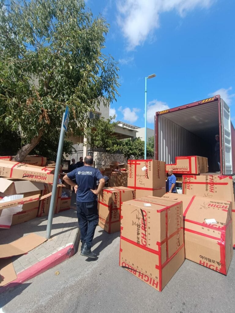 כמה קופסאות קרטון גדולות מפורקות ממכולה על המדרכה על ידי אנשים במדים, כחלק ממעבר דירה, תחת שמיים מעוננים חלקית. - דפנה פורמן, הרצאות, ייעוץ וטיפול רגשי במעברים וברילוקיישןש