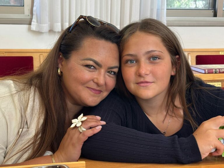 שתי נשים יושבות זו לצד זו ליד שולחן עץ, מחייכות למצלמה — אחת מהן חוגגת בבירור את יום הולדתה ה-13, והאמא שלידה קורנת מגאווה. ברקע נראים ספרים וכסאות עם כריות אדומות. - דפנה פורמן, הרצאות, ייעוץ וטיפול רגשי במעברים וברילוקיישןש