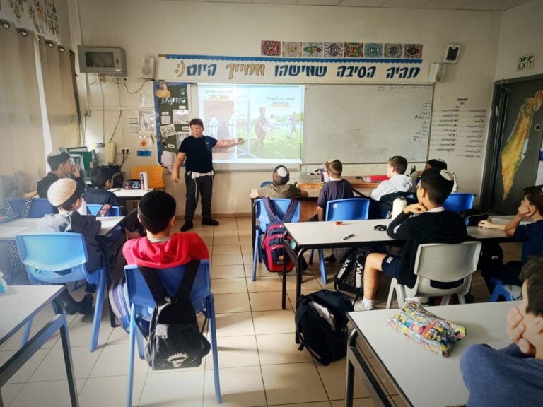 מורה עומד בקדמת הכיתה, מצביע על מסך ומנחה את הילדים בזמן שהם צופים ומקשיבים בקשב רב. - דפנה פורמן, הרצאות, ייעוץ וטיפול רגשי במעברים וברילוקיישןש