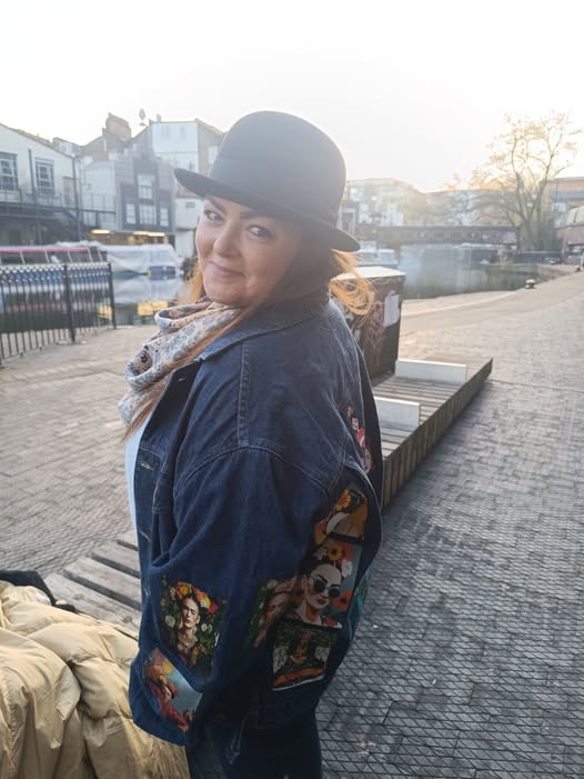 אישה חובשת כובע שחור, צעיף ומעיל ג'ינס מעוטר עומדת בחוץ ליד קו החוף על שביל מרוצף בשעת השקיעה, ומשקפת את נעם ותובנות ממסע הרילוקיישן שלה. - דפנה פורמן, הרצאות, ייעוץ וטיפול רגשי במעברים וברילוקיישןש