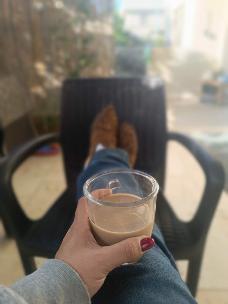 אדם עם לק אדום על הציפורניים מחזיק כוס קפה בידו, יושב עם רגליים מורמות על כיסא שחור בחוץ. - דפנה פורמן, הרצאות, ייעוץ וטיפול רגשי במעברים וברילוקיישןש