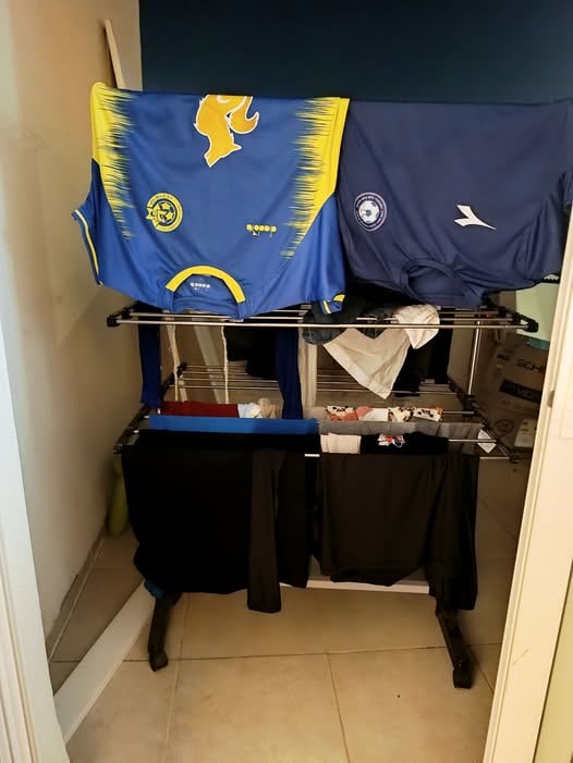 בגדים מתייבשים על מתלה שחור בתוך הבית, כולל גופיה ספורטיבית כחולה וצהובה, חולצה כחולה ובגדים כהים שונים. - דפנה פורמן, הרצאות, ייעוץ וטיפול רגשי במעברים וברילוקיישןש