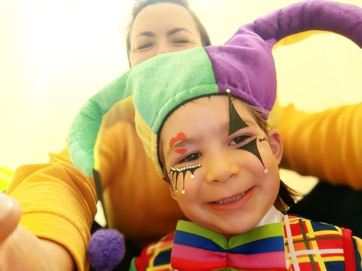 ילד לבוש בתחפושת ליצן צבעונית וצבוע בפנים מחייך בקדמת התמונה, חוגג את פורים ישראלי, ומאחוריו מבוגר מחייך. - דפנה פורמן, הרצאות, ייעוץ וטיפול רגשי במעברים וברילוקיישןש