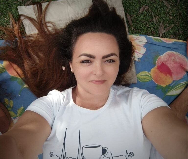 אישה עם שיער חום ארוך שוכבת על שמיכה בחוץ, מביטה במצלמה במבט נוסטלגי, ולובשת חולצת טי לבנה עם הדפס של כוס קפה. - דפנה פורמן, הרצאות, ייעוץ וטיפול רגשי במעברים וברילוקיישןש