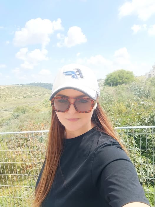 אישה עם שיער ארוך, חובשת משקפי שמש, כובע לבן וחולצה שחורה, עומדת בחוץ על רקע נוף ירוק ושמיים כחולים עם עננים. - דפנה פורמן, הרצאות, ייעוץ וטיפול רגשי במעברים וברילוקיישןש