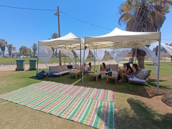 קבוצת אנשים יושבת תחת שני אוהלי סככה לבנים, עם שולחנות נמוכים וכריות על הדשא; מחצלת מעוטרת פרושה לפניהם, ועצי דקל נראים בסביבה — המקום המושלם למסיבות פרידה אינטימיות באווירה נינוחה תחת כיפת השמיים. - דפנה פורמן, הרצאות, ייעוץ וטיפול רגשי במעברים וברילוקיישןש