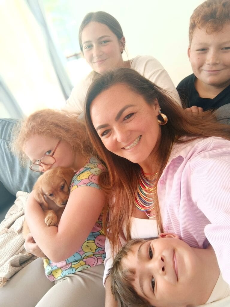 אישה יושבת על ספה עם ארבעה ילדים וכלבלב, כולם מחייכים למצלמה בחדר מואר היטב, בזמן שהם לוקחים הפסקה מ“לארוז את הבית” ומאריזת הקופסאות לקראת המכולה. - דפנה פורמן, הרצאות, ייעוץ וטיפול רגשי במעברים וברילוקיישןש