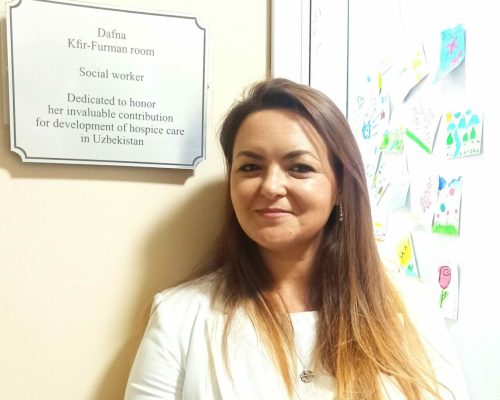 אישה עם ז'קט לבן עומדת מחייכת ליד שלט המכבד את תרומתה לפיתוח טיפולי ההוספיס באוזבקיסטן, ומדגישה את עבודתה המסורה עם ילדים חולי סרטן. - דפנה פורמן, הרצאות, ייעוץ וטיפול רגשי במעברים וברילוקיישןש